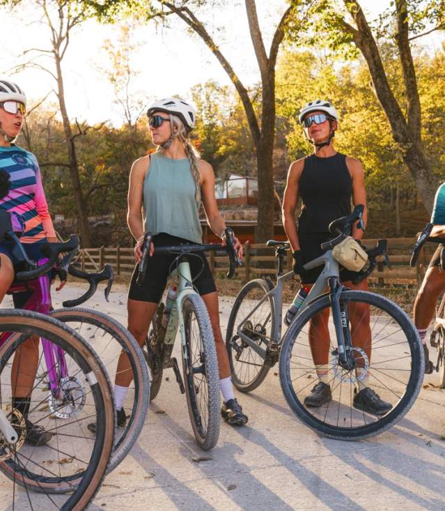 A group of five cyclists in helmets and athletic gear gather on a gravel path surrounded by trees, discussing their ride. Sunlight filters through the leaves.