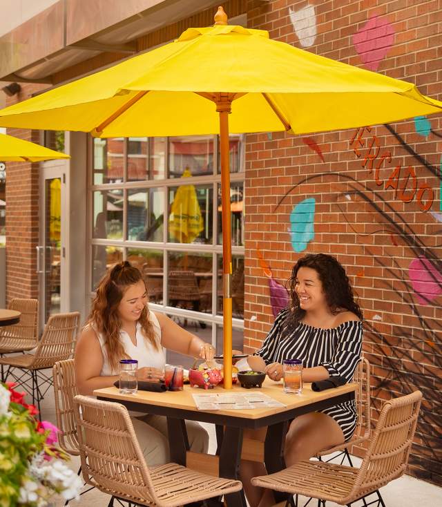 two women eating at an outdoor table at a restaurant sitting under a yellow umbrella with a mural on the wall behind them