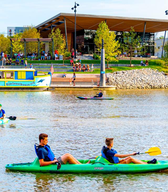 Kayakers enjoying a sunny afternoon on the river at Promenade Park with the Fort Wayne skyline and lush greenery in the background.
