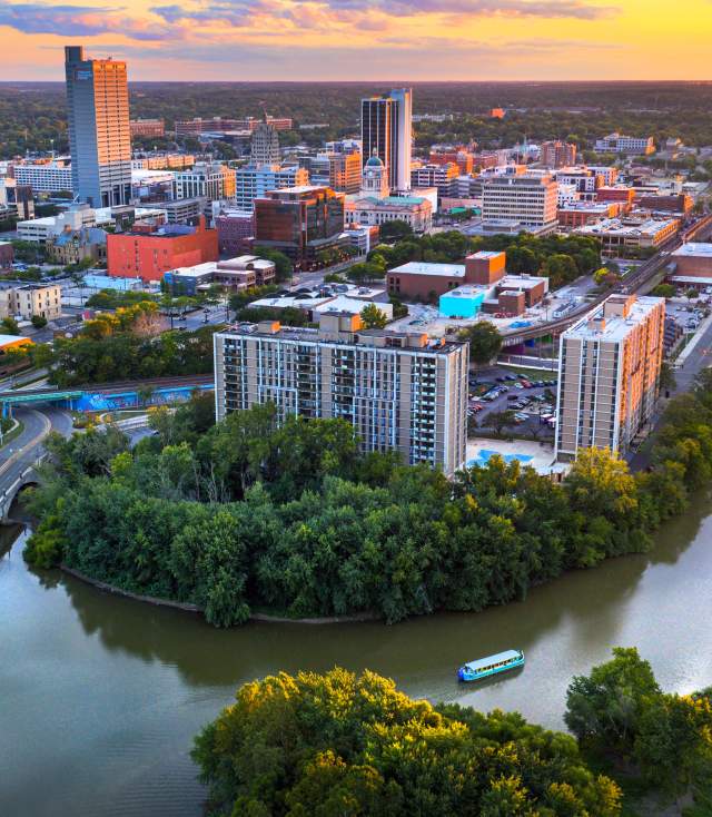 Drone shot of downtown Fort Wayne, Indiana at sunset with the confluence of the St Marys and St Joseph Rivers in the foreground and the skyline in the background. The Sweet Breeze canal boat is sailing on the St Marys River.