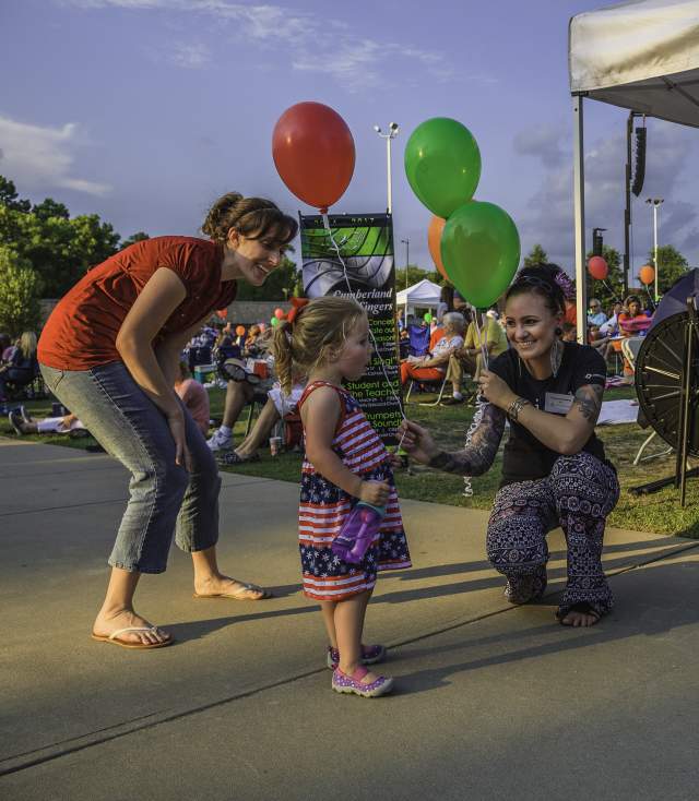 Toddler dancing on a sidewalk at an outdoor community event while adults smile and encourage her nearby