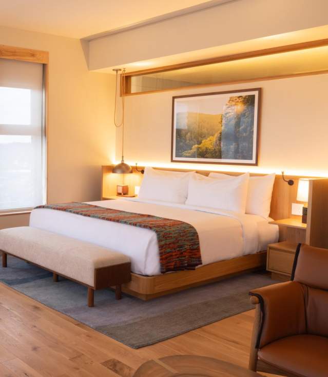 Modern hotel room featuring a king-sized bed, wooden furniture, and large windows with natural light. A small dining table and chairs are present.