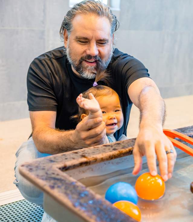 Father and young baby playing with ball in water feature at the Amazuem.