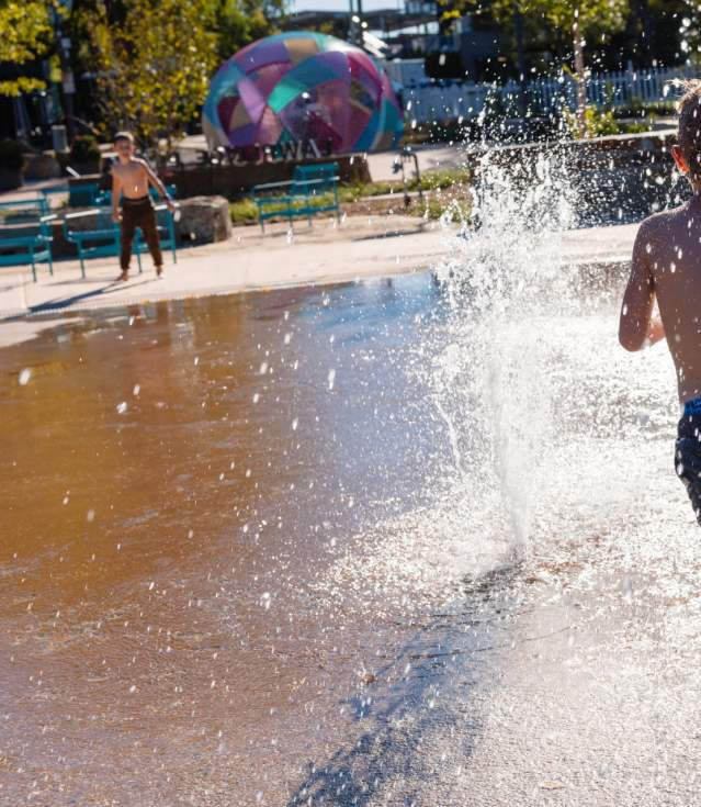 A young boy runs through a splash pad, water spraying around him. In the background, colorful structures and benches are visible.