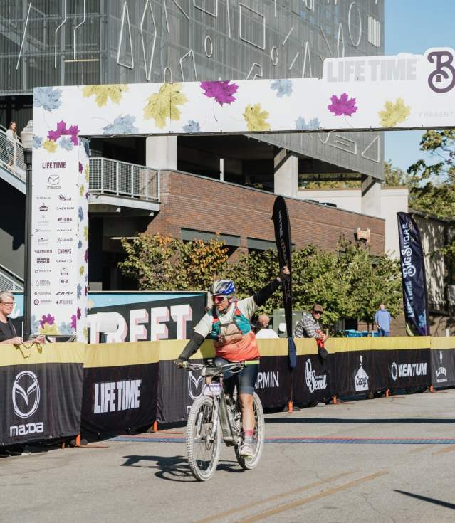 A cyclist crosses the finish line under a colorful archway at the Big Sugar Gravel event, surrounded by spectators and banners.