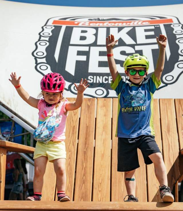 Two children throw their hands in the air like they just don't care and smile, from a large wooden chair at a bike festival, wearing helmets and colorful clothing, with the Bike Fest festival banner in the background.