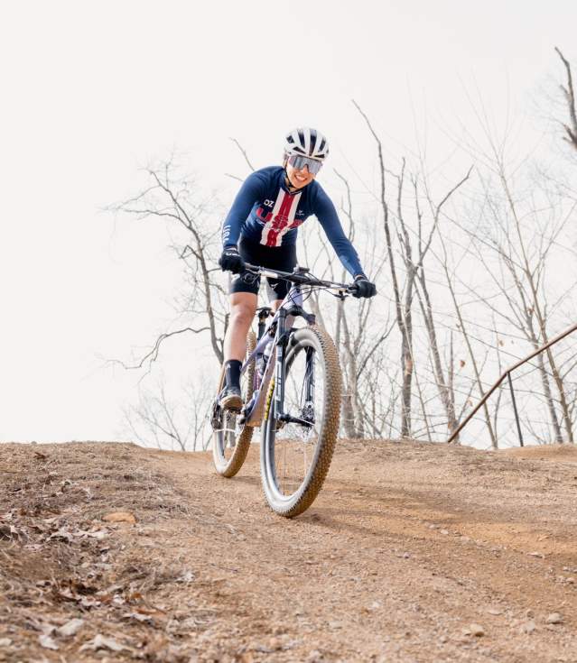 A cyclist navigates a dirt trail, riding downhill on a mountain bike surrounded by bare trees and a clear sky. The scene captures an active outdoor adventure.