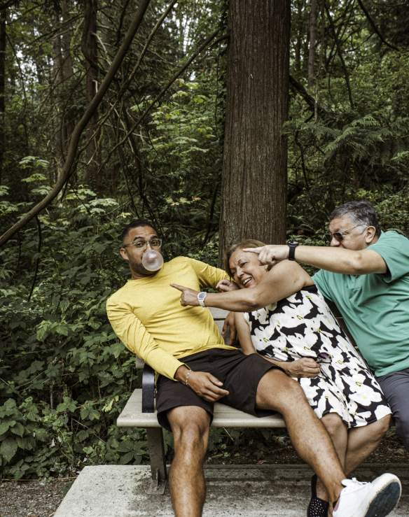 A man in a yellow shirt blows a bubblegum bubble while sitting on a forest park bench as an older couple beside him laugh and reach across playfully to try to pop it.