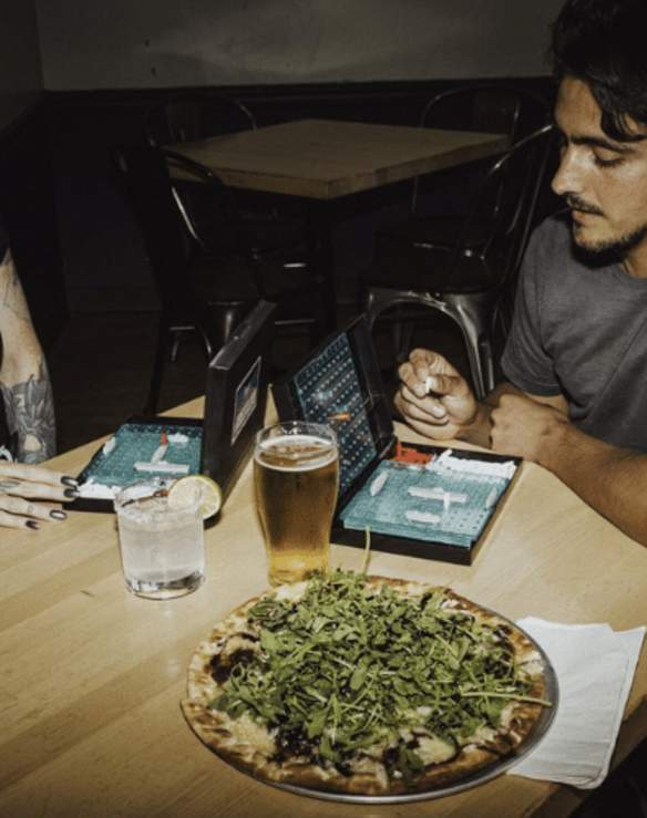 Two people playing battleship at a restaurant with pizza.
