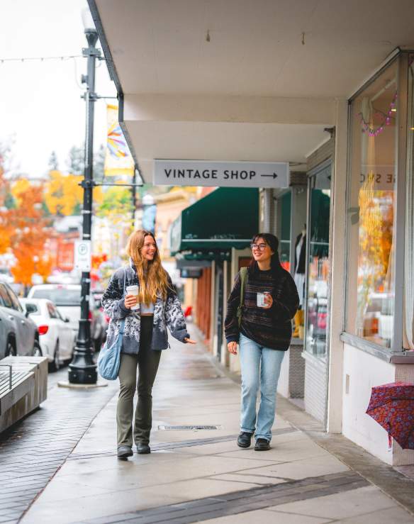 Two people stroll past shops on a rainy autumn day in downtown Nanaimo, holding coffee as colourful fall trees line the street.