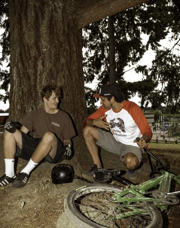 Two men sitting leaning on a tree with a bike.