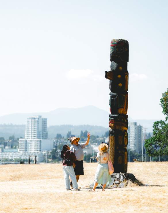 Three people stand at the base of a totem pole on Saysutshun with Nanaimo’s skyline visible across the harbour in the background.