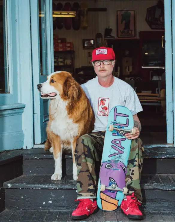 Aa man and his dog sitting with a skateboard.