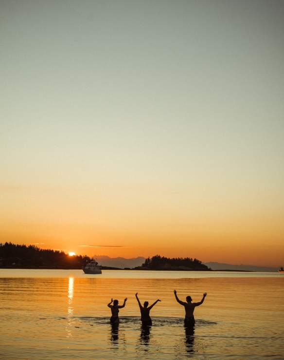 Three people standing in calm ocean water at sunset with arms raised.