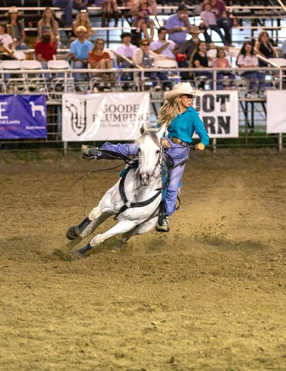 Woman in a Teal shirt Barrel Racing with a White Horse.