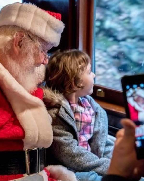 Santa sits next to a little girl as both look out the window on a Colebrookdale Railroad holiday excursion. A person's hand, holding a cellphone, is to the far right of the screen, capturing the picture of Santa and the little girl.