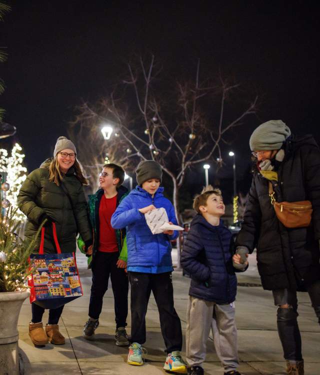 A group of two adults and three children walk together on a decorated city sidewalk at night during the holiday season. They are bundled in winter coats, hats, and gloves. Festive lights and small decorated trees line the shop windows on the left. One adult carries a colorful tote bag, and one child holds a snack. Everyone appears cheerful and engaged with one another as they enjoy the winter evening.”