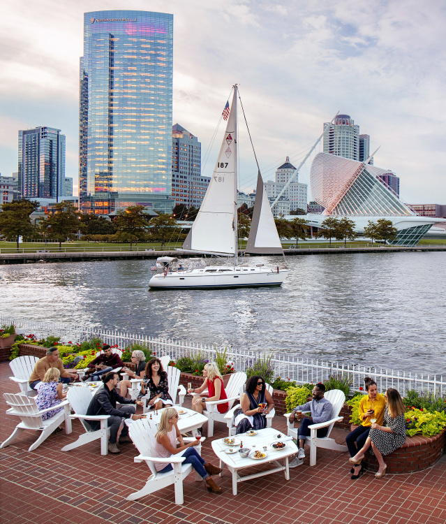 a group of people eating on the patio in front of the Milwaukee skyline with a sailboat in the harbor