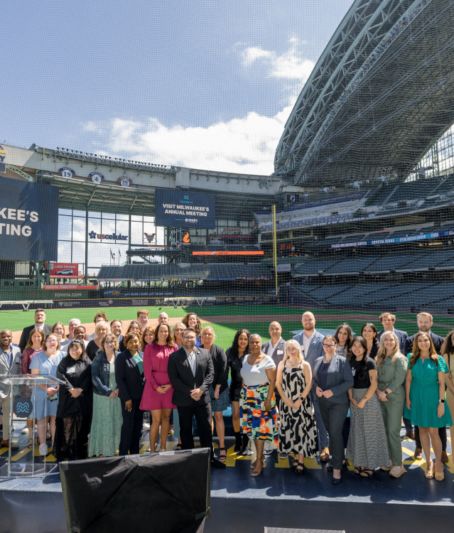 Group photo of attendees at Visit Milwaukee's Annual Meeting, posed on a stage at American Family Field (Milwaukee Brewers stadium) with stadium seating and banners visible in the background.