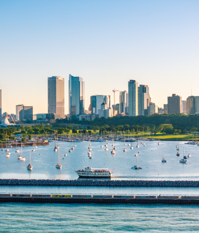 A wide view of Milwaukee’s lakefront skyline on a clear day, with dozens of sailboats anchored in a calm marina and a tour boat cruising along the breakwater in the foreground. Green parkland lines the waterfront beneath a cluster of downtown high-rise buildings and glass towers.