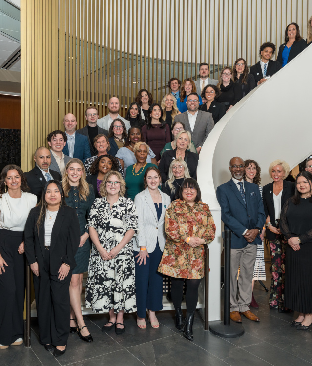 Large group of professionals posing on a curved staircase inside a modern building with wood slat walls and glass railings.