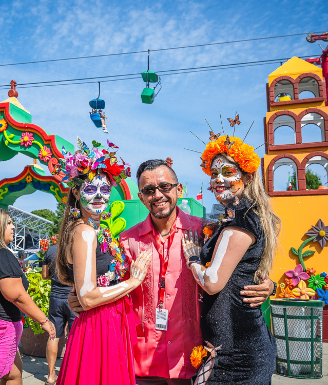 Three people pose and smile at a vibrant outdoor festival in Milwaukee. Two wear colorful Day of the Dead-style face paint and flower crowns, standing next to a man in sunglasses. Behind them is a festive archway and colorful decorations, with people enjoying the event under a bright blue sky.