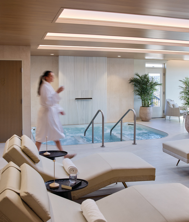 A tranquil spa lounge features cushioned reclining chairs, rolled towels, and a bubbling hydrotherapy pool beneath warm wood-paneled ceilings. A guest in a robe walks through the bright, modern space, creating a relaxing atmosphere focused on wellness and rejuvenation.