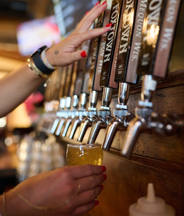 Close-up of a bartender pouring a golden beer from a row of draft taps labeled with Milwaukee Brewing Company beers into a small tasting glass.