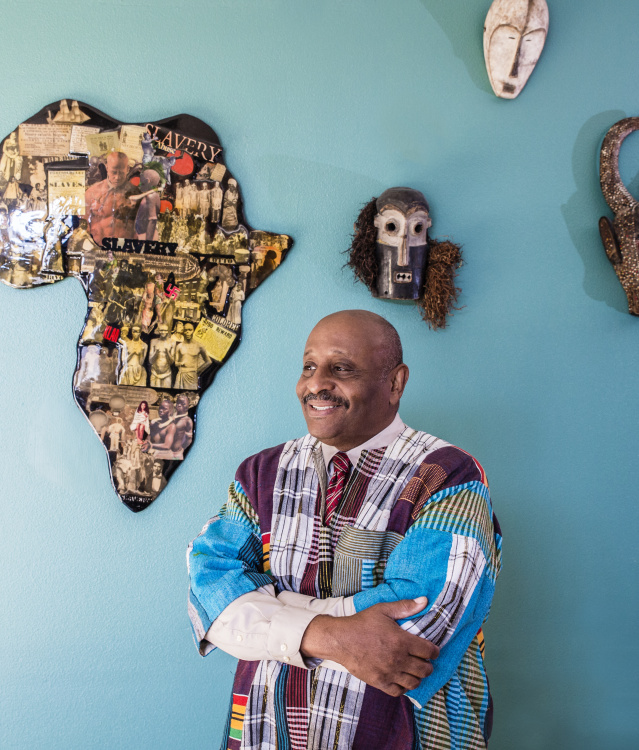 black man in traditional dress standing in front of a teal wall with a map of Africa