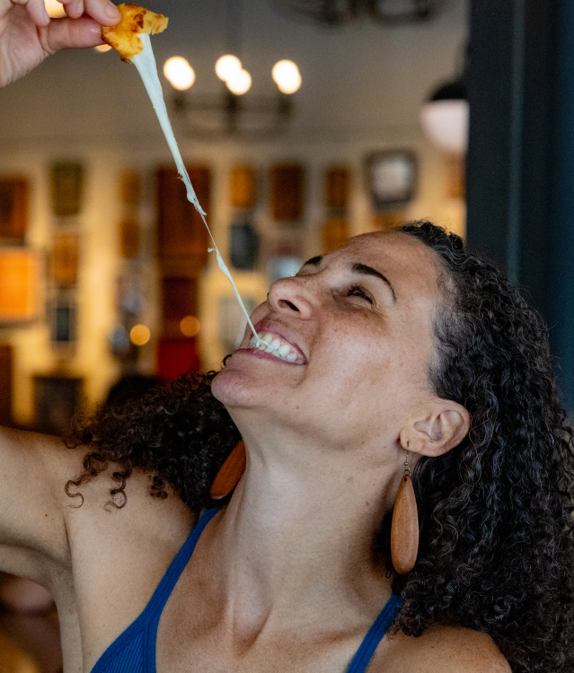 A smiling woman pulls apart a cheesy appetizer, creating a long stretch of melted cheese as she enjoys a meal inside a warmly lit restaurant. The background is softly blurred with artwork and lights visible.