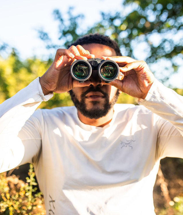 A man in a white long-sleeved shirt holds binoculars up to his eyes while standing outdoors in a sunny, tree-filled setting with green foliage and dappled sunlight in the background.