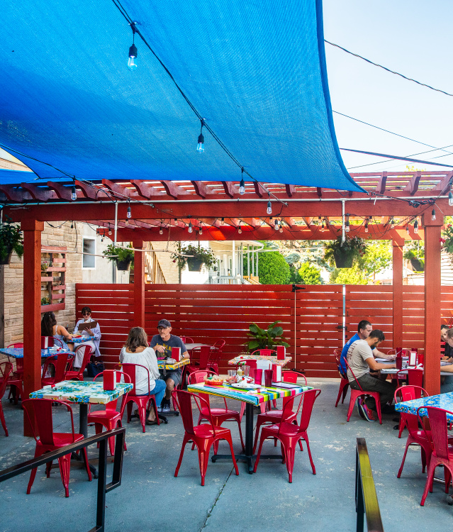 Outdoor patio dining area at Café Corazón featuring red chairs, colorful tablecloths, and hanging plants under a shaded pergola with a bright blue canopy. Diners are enjoying meals in a relaxed, vibrant setting.