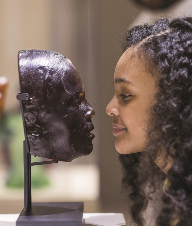 A young visitor leans in to closely examine a glass face sculpture displayed on a stand at a museum. The side profiles of the sculpture and the visitor align, creating a striking visual comparison in the gallery setting.