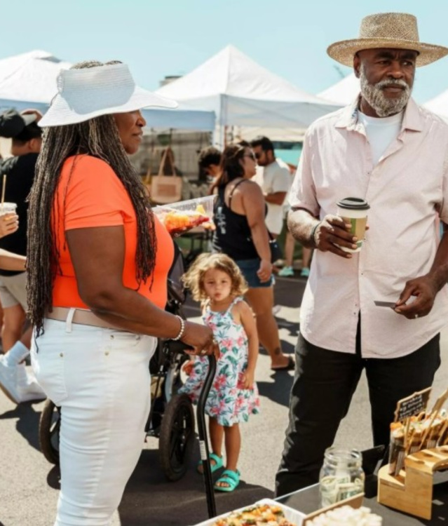 Visitors browse vendor booths at an outdoor market on a sunny day. Two people chat while holding coffee near a handcrafted goods display, as families and shoppers explore the bustling festival in the background.