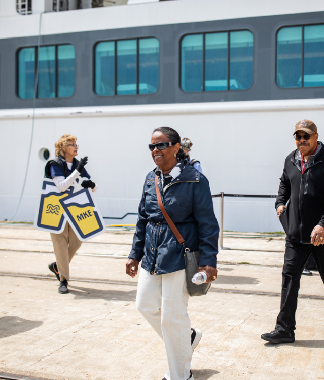 Visitors walk along a dock beside a large cruise ship, greeted by people holding Milwaukee-themed signs, creating a welcoming arrival scene on the waterfront.