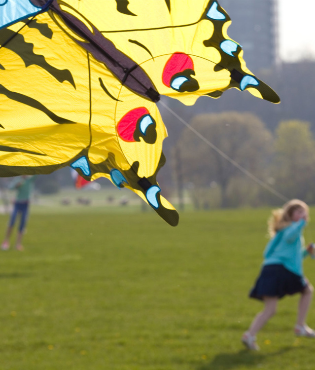 A young girl running into the distance holding a string attached to a kite shaped like a butterfly with yellow coloring on the body and black stripes and blue accents throughout the edges, all will bright green grass and the city skyline in the background.