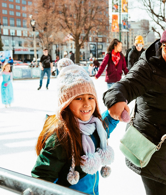 a little girl ice skating on a public ice rink while holding her mom's hand