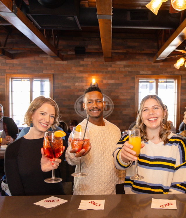 “A group of six smiling adults stand at a bar, holding up cocktails in a celebratory toast. They are positioned side by side in a warmly lit space with brick walls and exposed beams. Each person is looking toward the camera, and various drinks in bright colors—red, orange, and amber—are visible in their hands. Bar napkins and branded coasters sit on the counter in front of them.