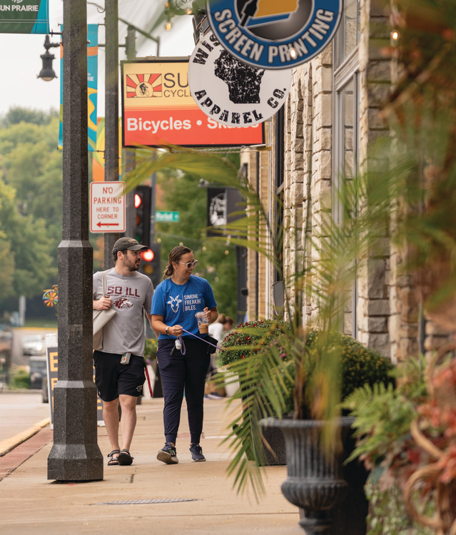 Two people stroll along a lively downtown sidewalk lined with local shops, hanging business signs, and colorful planters. Decorative lights and welcome banners add to the inviting streetscape as they explore the district on a warm day.
