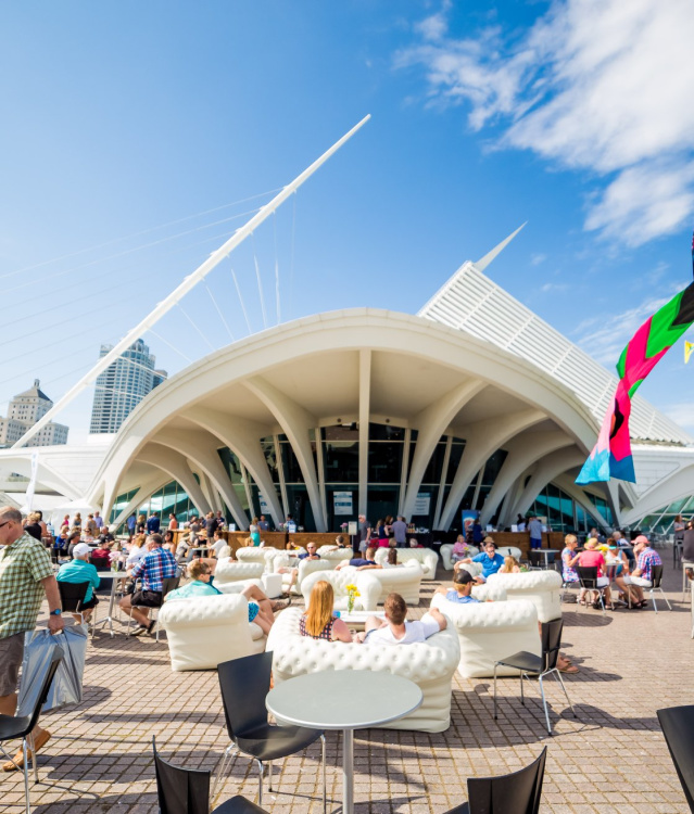 A wide angle shot of the Milwaukee Art Museum with festival goers sitting on the patio outside, with flags blowing in the wind against a beautiful blue sky.