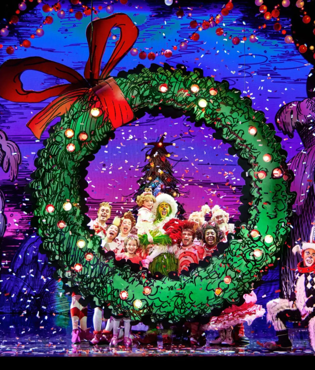 A colorful stage scene from a Christmas-themed musical featuring characters in bright costumes gathered around a giant green wreath with a red bow. Snow-like confetti falls from above, and whimsical, snow-covered houses are visible in the background.