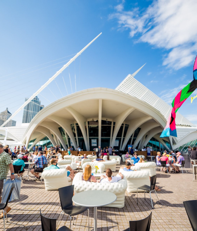 A wide angle shot of the Milwaukee Art Museum with festival goers sitting on the patio outside, with flags blowing in the wind against a beautiful blue sky.