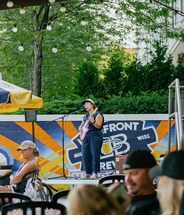 A woman in a navy blue romper singing into a microphone on a stage, holding a pink guitar, with people in front of her sitting at tables under gold and blue umbrellas on a patio along the riverfront with trees in the background.