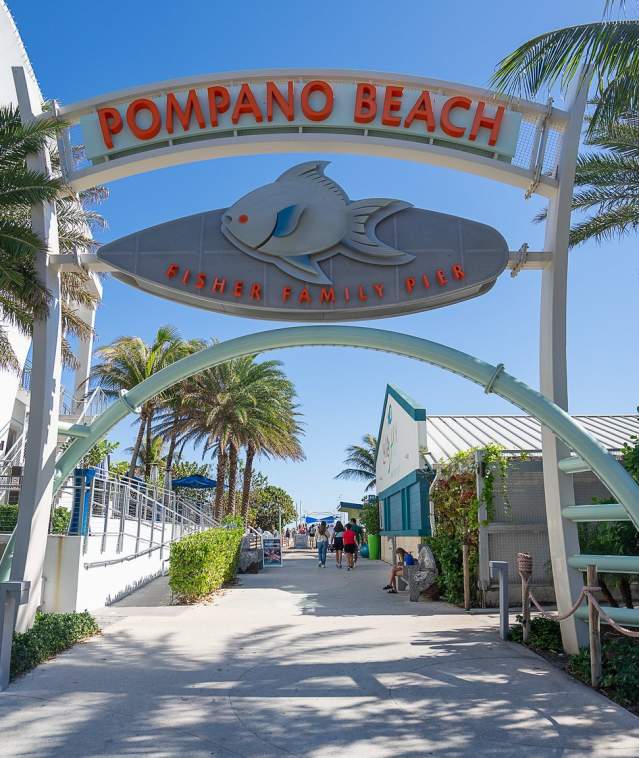 An archway above a wide entrance walkway with palm trees on both sides. The sky is bright blue, and the sun is casting shadows of the palm trees on the walkway. Above the arch is a sign that says, "Pompano Beach, Fisher Family Pier."
