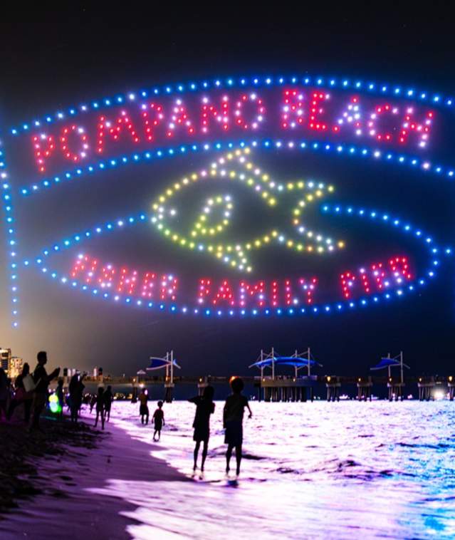 The Pompano Beach Fisher Family Pier Entrance sign depicted by drones in the night sky at the beach in Pompano Beach, FL