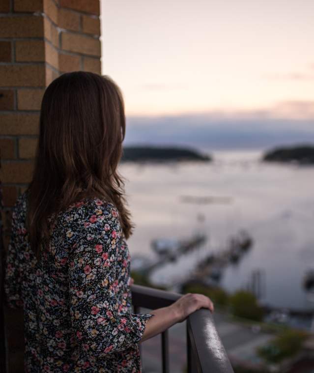 A person stands on a balcony overlooking a harbour with boats and distant islands at sunset.