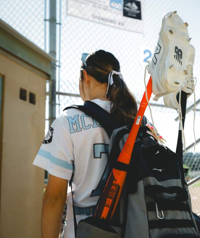 A softball player in uniform walks toward a field, carrying a bat and cleats attached to their backpack.