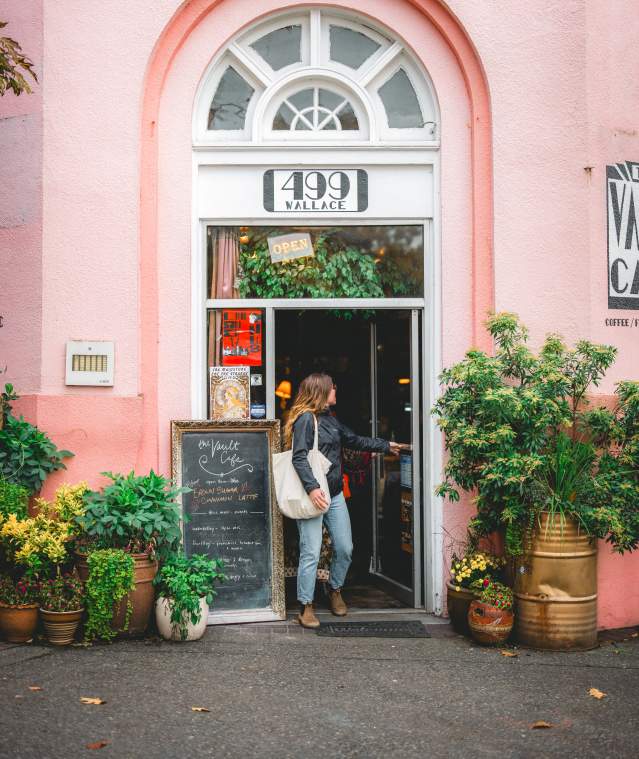 A person opens the door to a café with a pink exterior, surrounded by potted plants and a chalkboard sign.