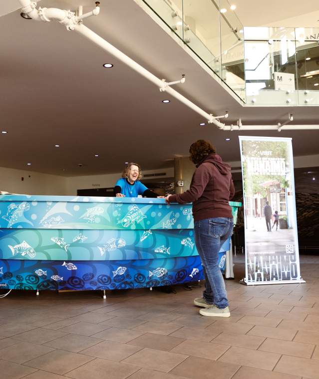 A visitor speaks with staff at a tourism information desk inside the Nanaimo Museum, with promotional displays nearby.