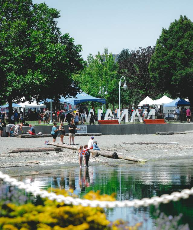 People gather along a waterfront park in Nanaimo (Maffeo Sutton Park), with children wading in the water and market tents and the ‘Nanaimo’ sign in the background.